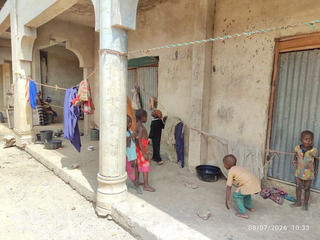 Children play and do chores outside a building with clothes hanging on lines. Two people sit in the shaded area.