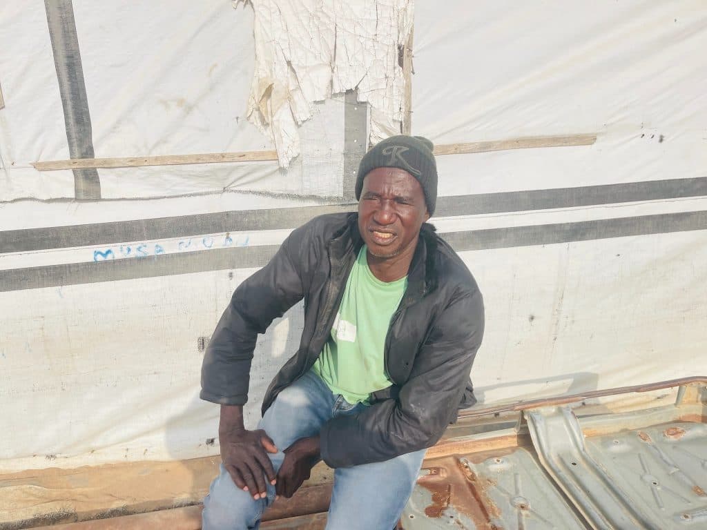 A person in a jacket and beanie sits on a metal surface against a tent-like background outdoors.