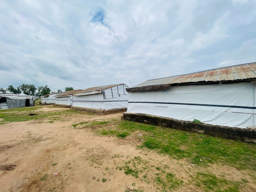 Corrugated metal and tarp shelters under a cloudy sky, surrounded by sparse grass and dirt.