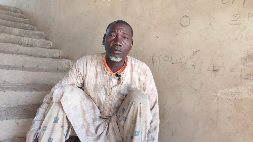 A man in patterned clothing sits on stairs against a wall with faint markings.