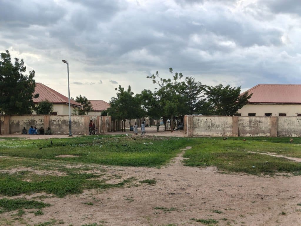 People sitting and standing near a walled area with trees and cloudy sky in the background.