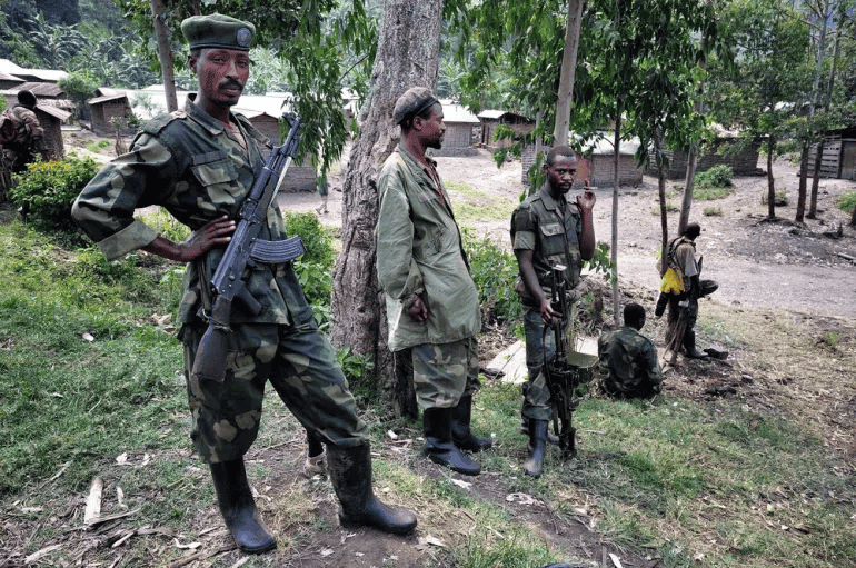 Soldiers in camouflage stand with rifles under trees in a rural setting with wooden buildings in the background.
