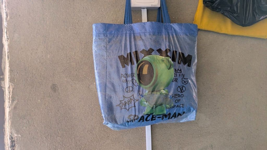 Blue tote bag with a cartoon green spaceman design and text, hung on a wall.