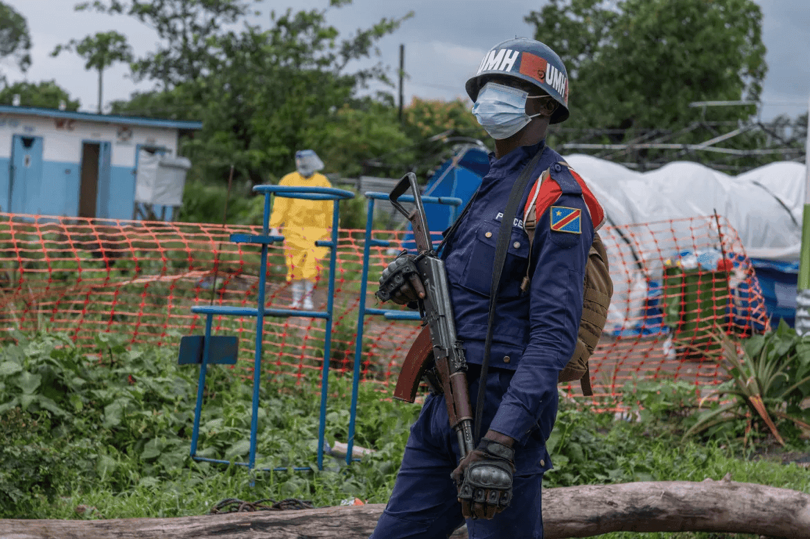 A masked peacekeeper stands with a rifle in a fenced area, while a person in protective gear walks in the background.