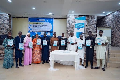 Group of people at a book launch event, each holding a copy of the book. Banners and a table with a white cloth are in the background.