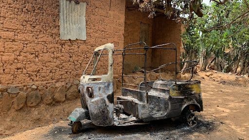 Burned-out auto rickshaw with no roof sits on dirt ground near a mud-brick building under a tree.