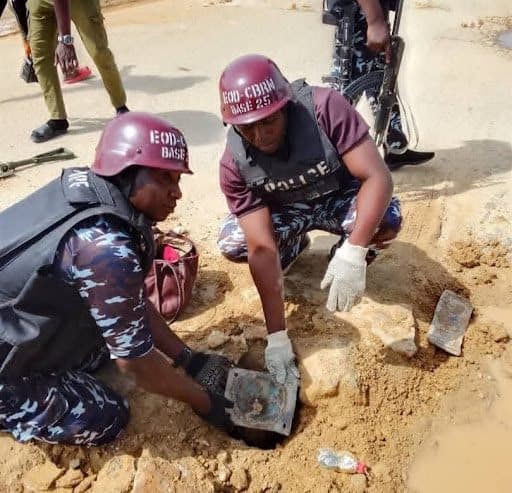 Two uniformed individuals examining an object in a small excavation, wearing protective helmets and gloves, with a third person in the background.