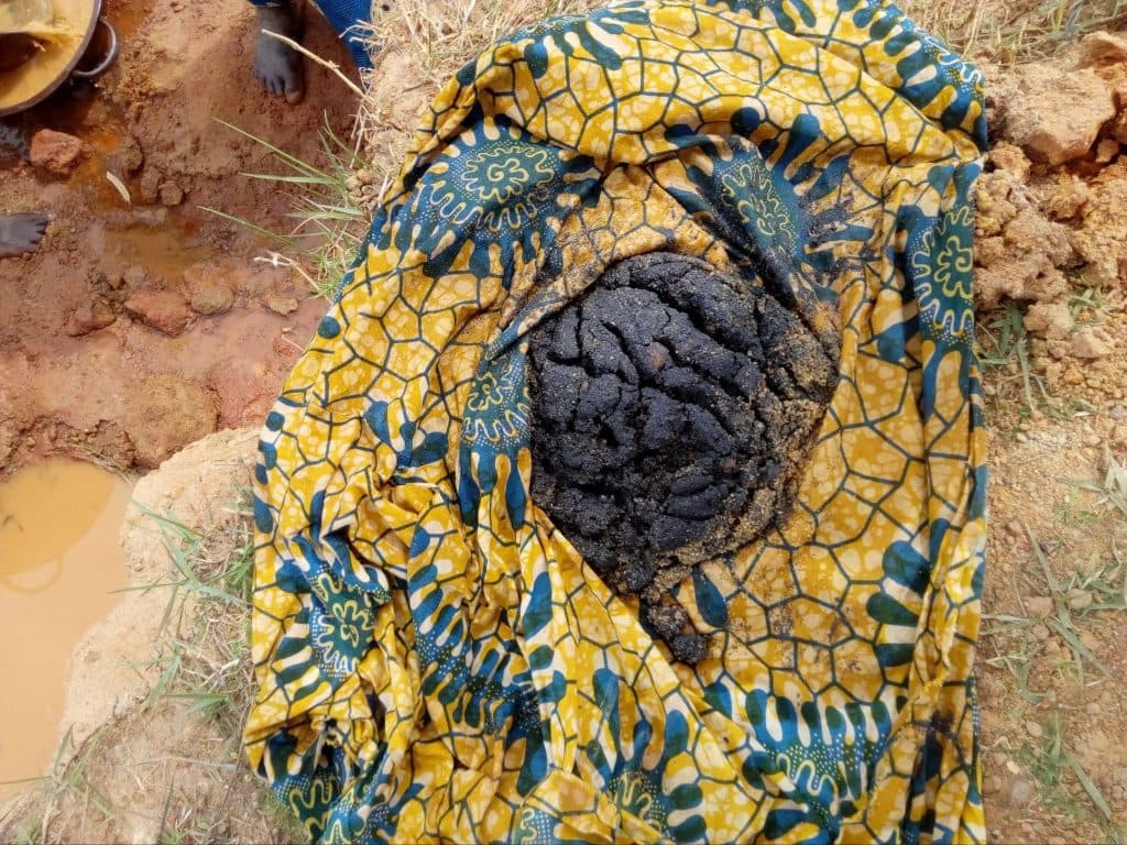 A textured black disc rests on a colorful patterned cloth, surrounded by earthy ground, grass, and a muddy water hole.