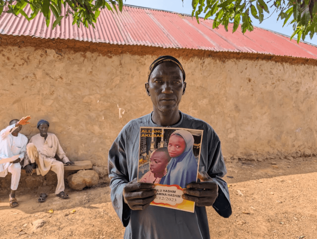Man holding a photograph of two children outside; three people sit on a bench in the background.