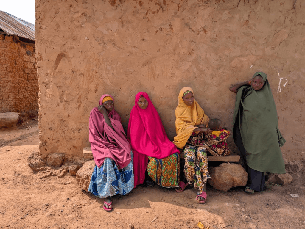 Four children in colorful clothing sit against a textured stone wall in a rural setting.