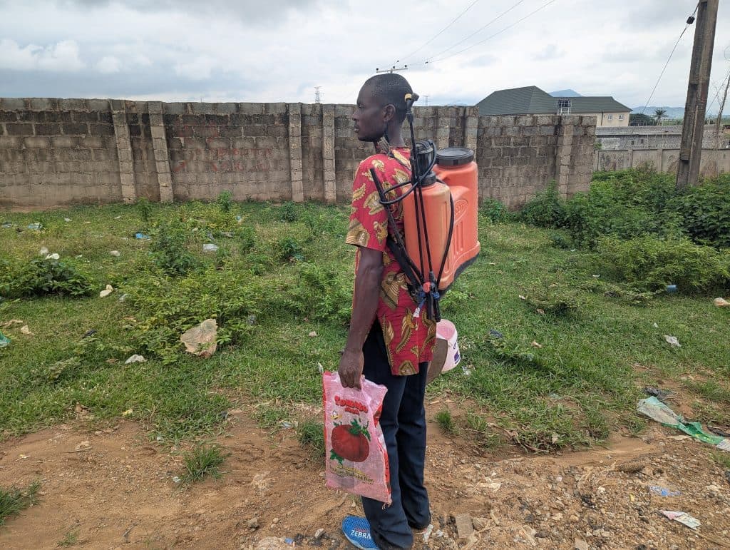 Person standing on grass, wearing a backpack sprayer and holding a bag, with a brick wall and houses in the background.