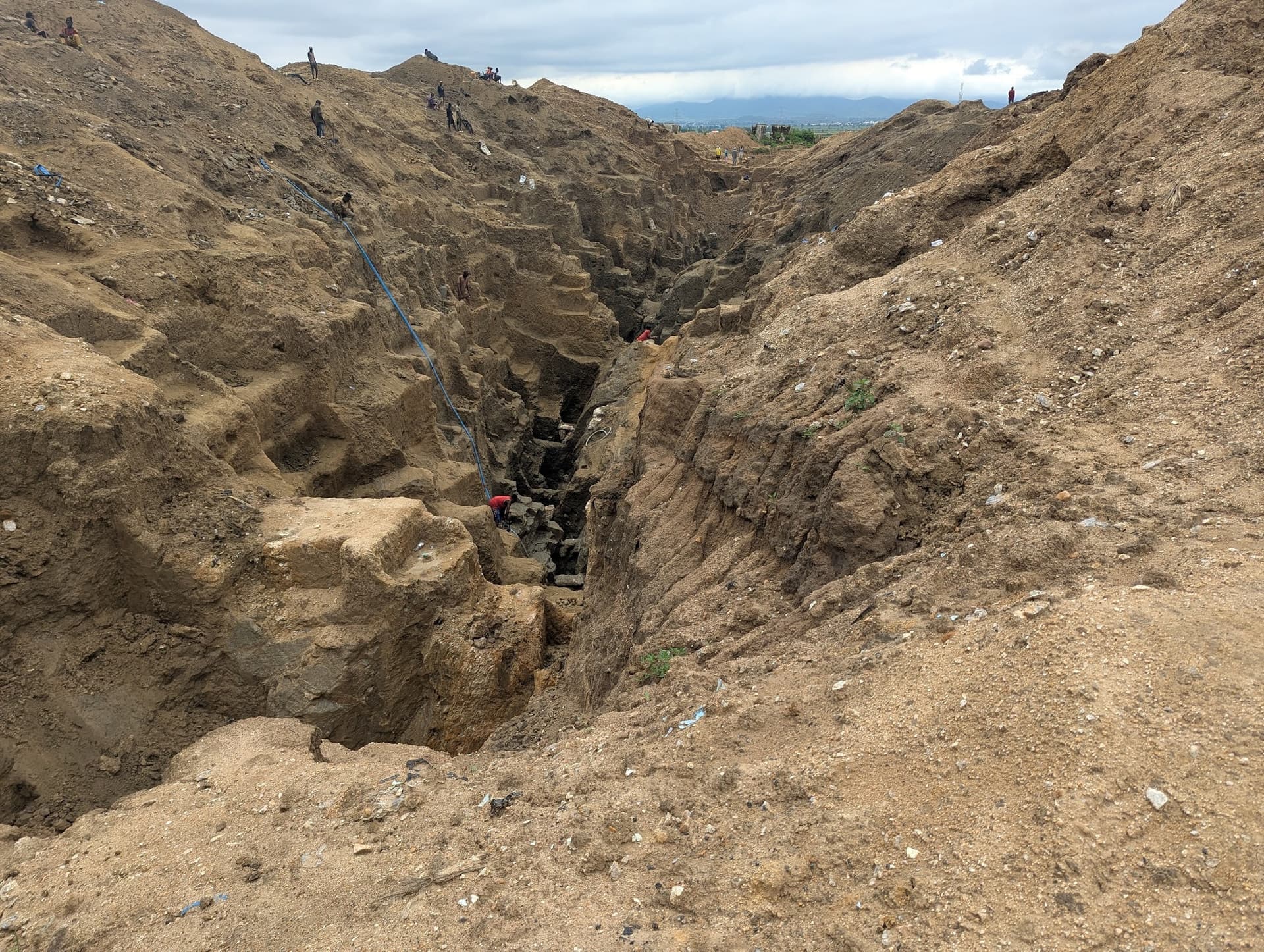 Rugged landscape with deep trenches and scattered workers climbing steep dirt paths under a cloudy sky.
