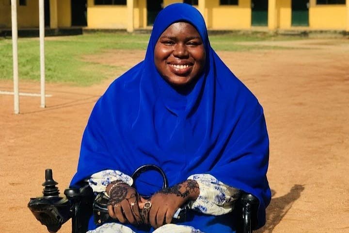 Smiling person in a blue hijab sitting in a wheelchair outdoors with a sunny yellow building in the background.