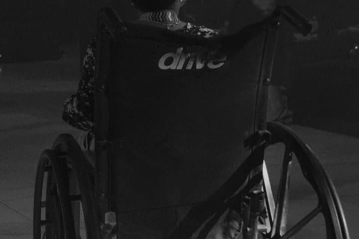 Back view of a person sitting in a wheelchair with "drive" on the backrest, wearing a patterned top, in a dimly lit setting.