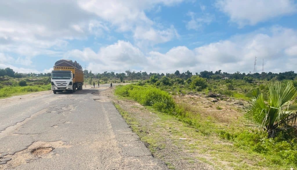 A truck on a rural road with potholes, surrounded by greenery under a bright, partly cloudy sky.
