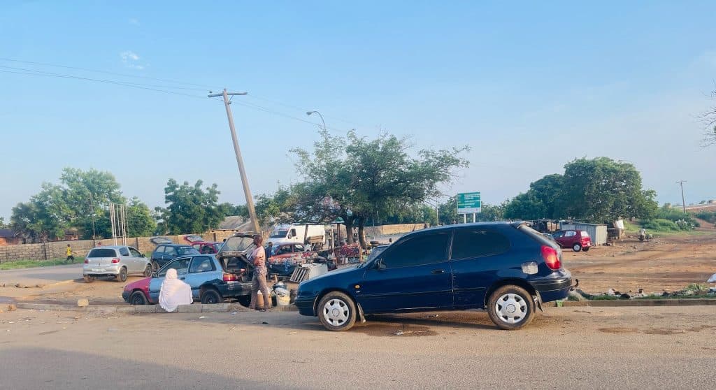 Street scene with cars parked on the roadside. People are gathered near a car with its hood open. Trees and a blue sky in the background.
