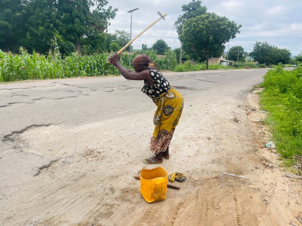 Person repairs roadside with a wooden tool near a lush green landscape and cornfield under a cloudy sky.