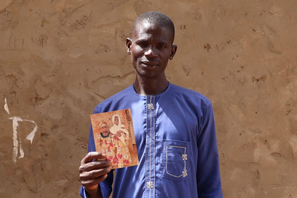 Man in blue shirt holding a photograph of two people, standing against a textured wall background.