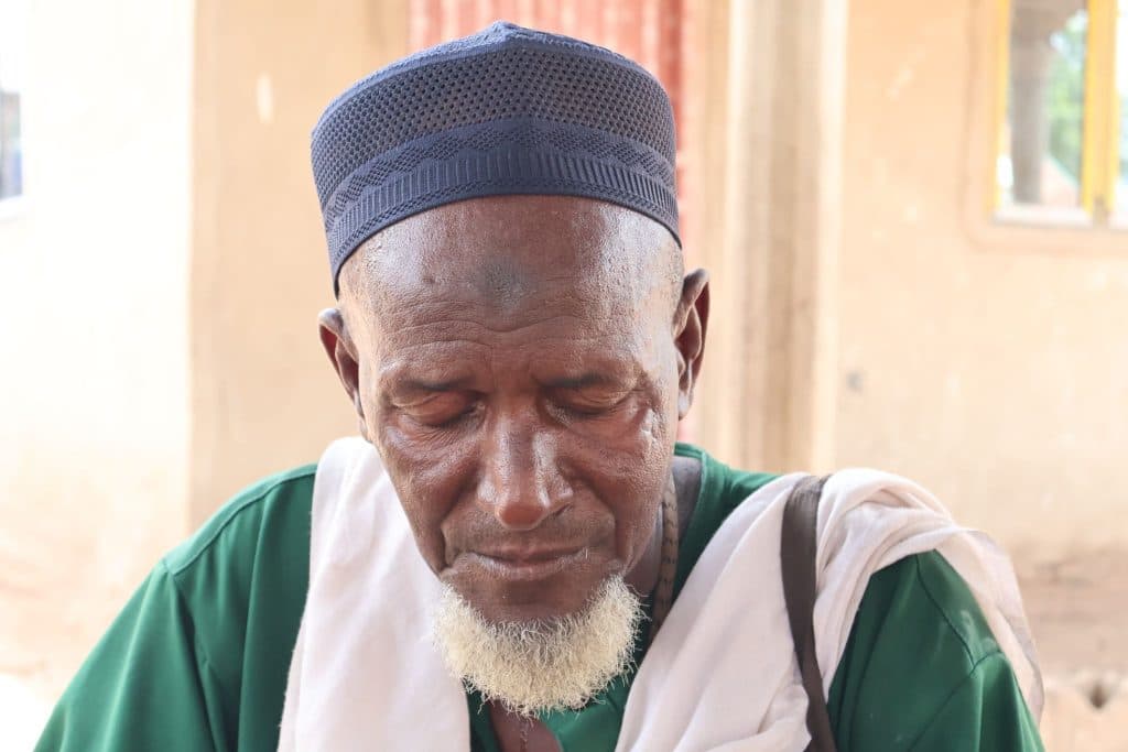 Elderly man with a white beard and closed eyes, wearing a dark cap and green garment.