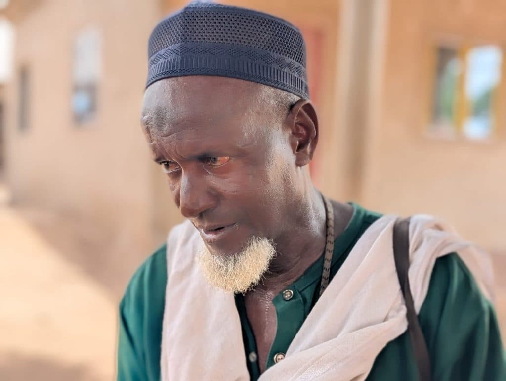 Elderly man with a white beard wearing a navy cap and green shirt outdoors, looking to the side.