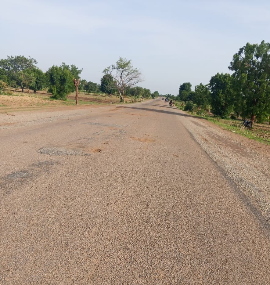 Wide, empty rural road with scattered potholes, bordered by trees and grass under a clear sky.