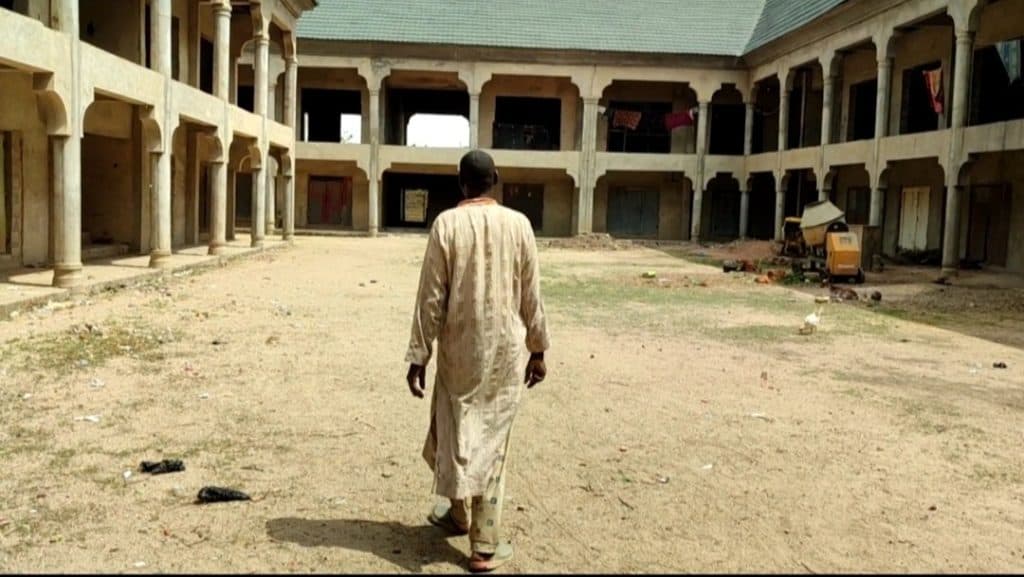 Person walking in a sandy courtyard surrounded by a two-story building with arches.