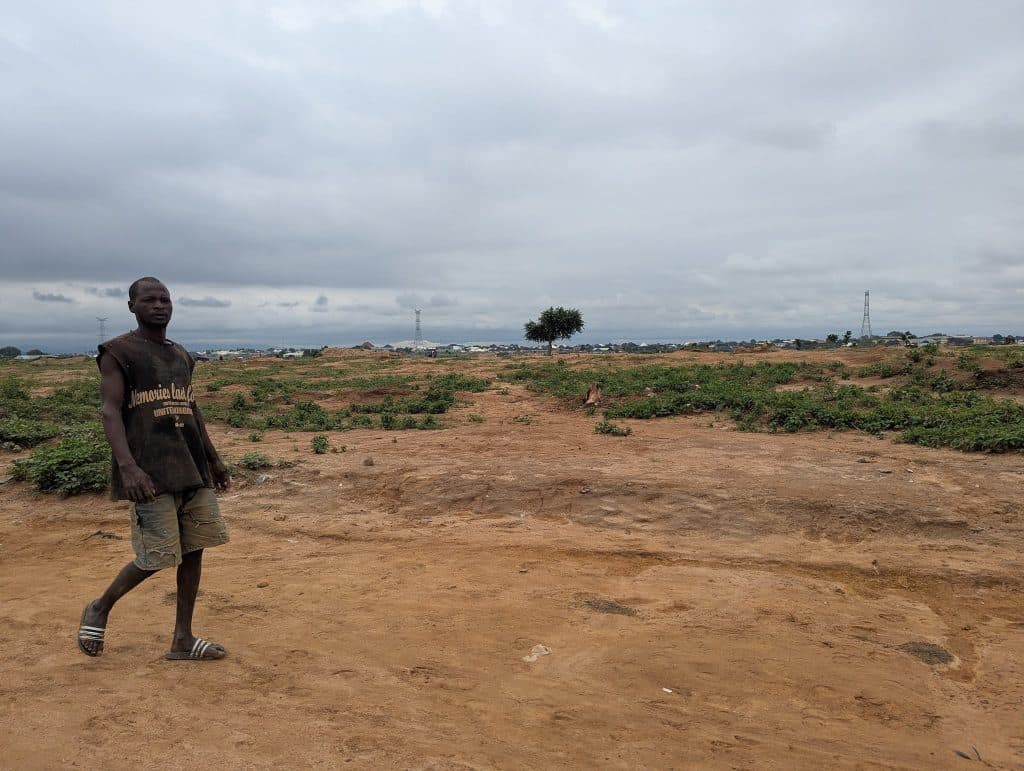 Man walking on a sandy, open field under a cloudy sky, with a distant tree and city skyline in the background.