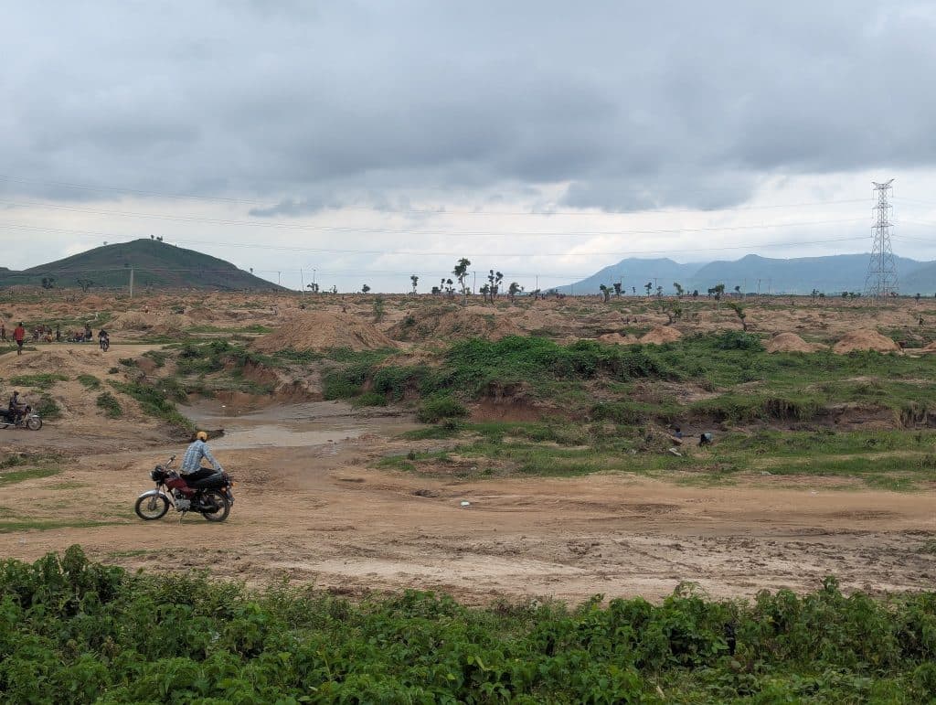 Motorcyclist rides on a dirt path through a vast, green landscape with hills and cloudy skies in the background.