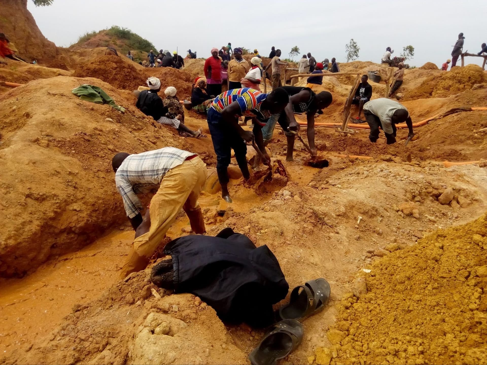 People working in a sandy area, digging and searching through the soil. Scattered tools and a pair of sandals are visible.