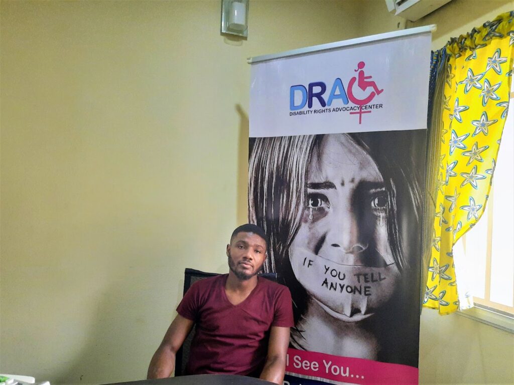 Idris Agboluaje wears a short-sleeve, plain red t-shirt and sits in front of a roll-up banner for the Disability Rights Advocacy Centre.