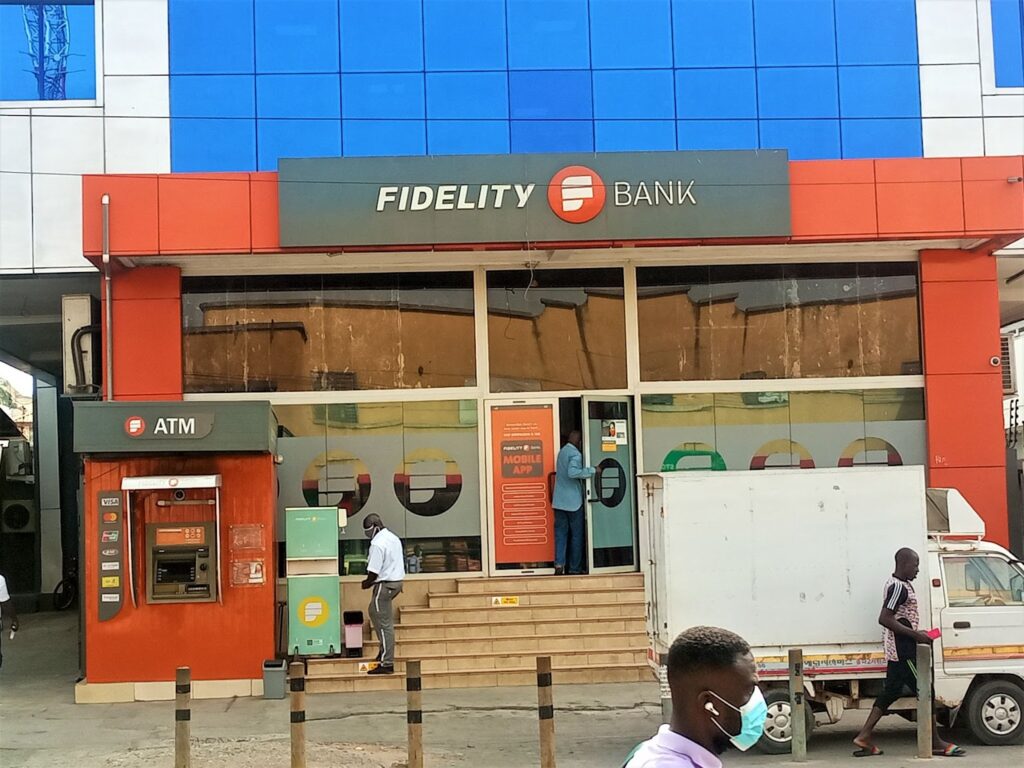 A tall staircase with seven steps leads into the main entrance of the Fidelity Bank branch at the Kwame Nkrumah Avenue in Ghana. The building is covered in red, blue, and silver tiles. There is an ATM outside on the ground floor.