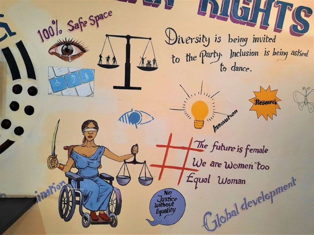 Wall art at the Abuja office of the Disability Rights Advocacy Centre. There is a painting of a blindfolded Lady Justice holding the scale and a sword and sitting on a wheelchair. There are other paintings too, of a lightbulb, a disability-friendly keyboard, a teary eye, and a hashtag. There are words in the painting including those that say "diversity is being invited to the party. Inclusion is being asked to dance" and "the future is female. we are women too. equal women."