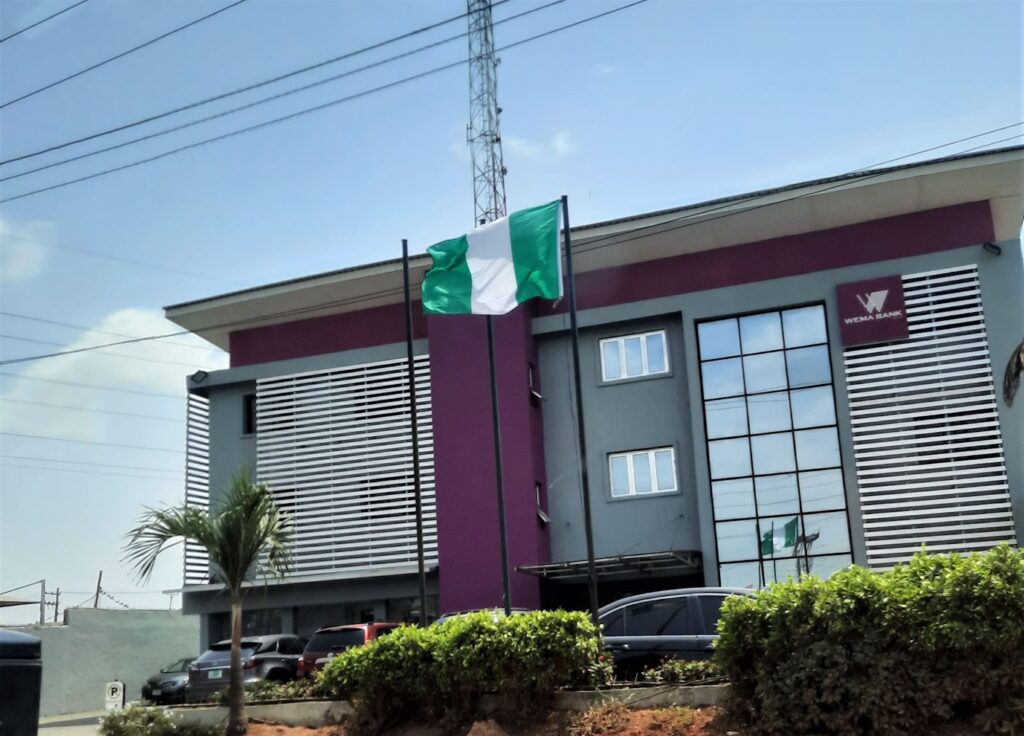 The picture shows outside one branch of Wema Bank in Lagos, Nigeria. The walls are painted purple. A Nigerian flag is flying by the roadside. Cars are parked outside the building.