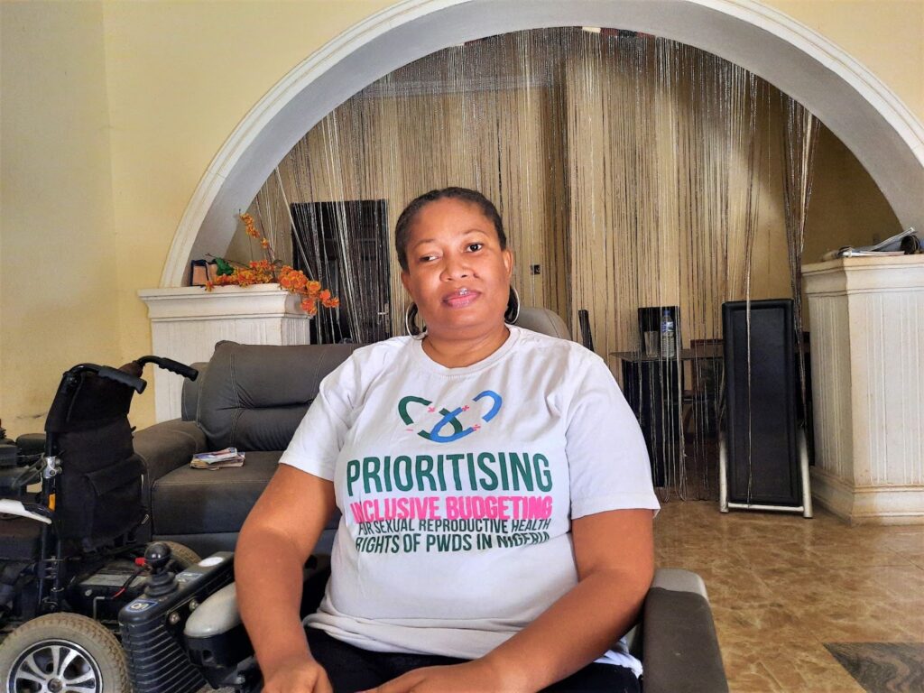 Abdulrahman Violet is in the living room of her apartment in Abuja. She is looking straight at the camera. She is sitting on a motorised wheelchair. She is wearing a white t-shirt that says, "prioritising inclusive budgeting for sexual reproductive health rights of persons with disabilities in Nigeria."