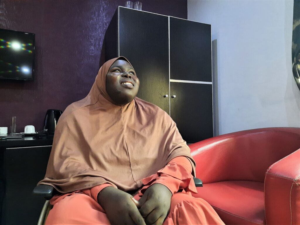 Fatimah Aderohunmu is smiling in this picture and looking towards the ceiling. She is in a hotel room for the interview and sits on a wheelchair. She is wearing a pink dress and a brown hijab.