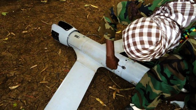 A person in a camouflage uniform examining an unmanned aerial vehicle (UAV) on the ground.