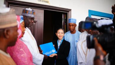 A group of dignitaries with a woman holding a UNICEF folder by a building's main entrance, with cameras pointing at them.