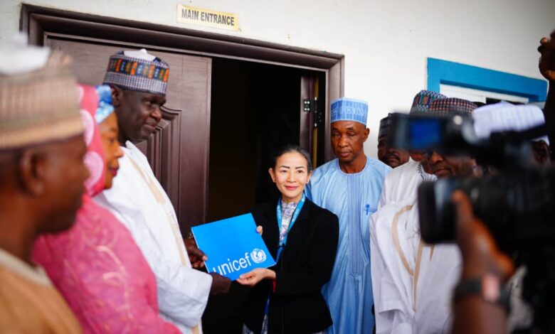 A group of dignitaries with a woman holding a UNICEF folder by a building's main entrance, with cameras pointing at them.
