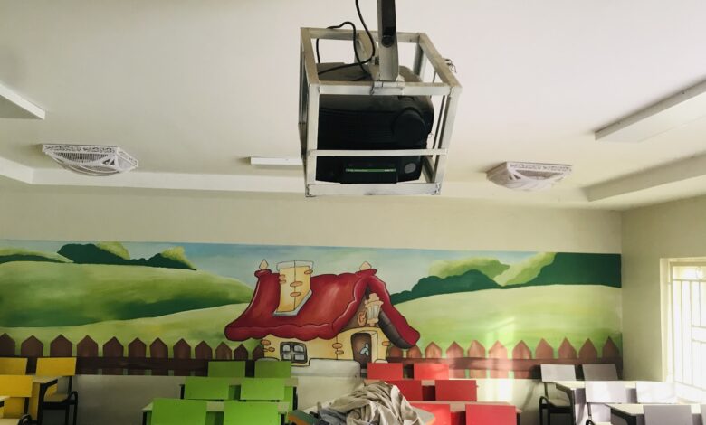 Classroom with colorful desks, a mural of hills and a house on the wall, and a projector hanging from the ceiling.