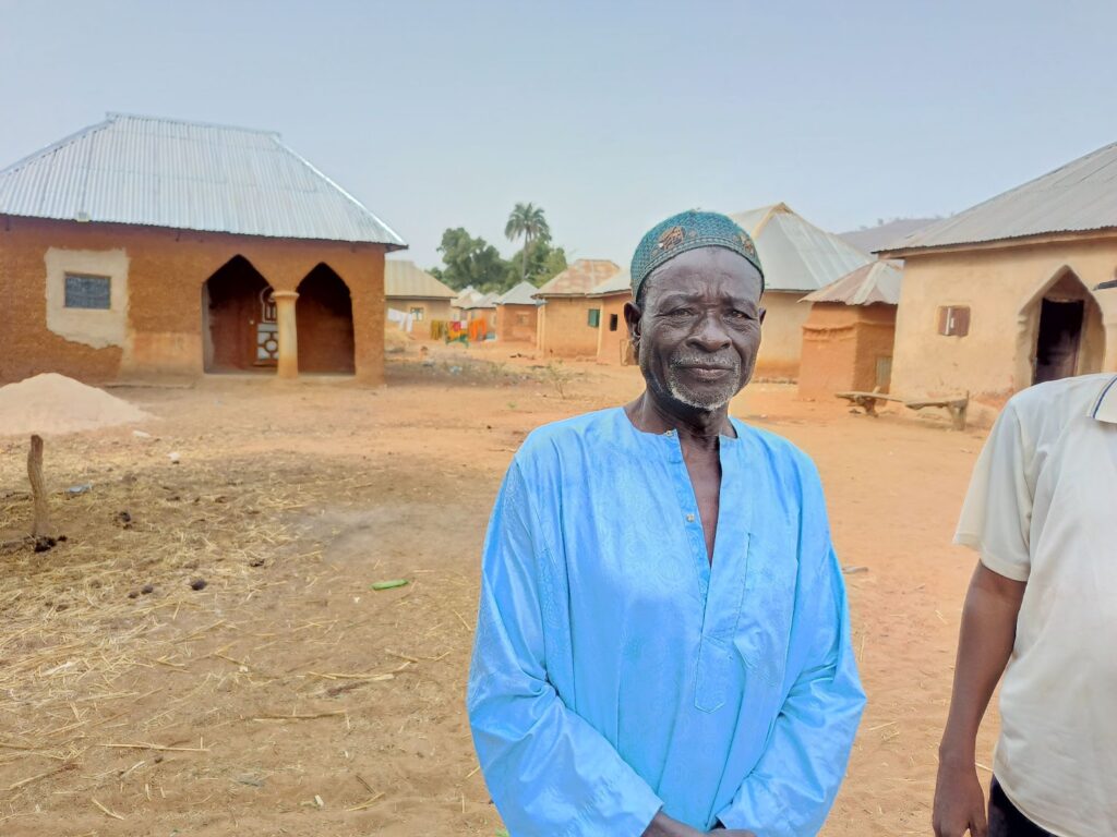 Elderly man in a blue shirt with a cap stands before rural African village homes.