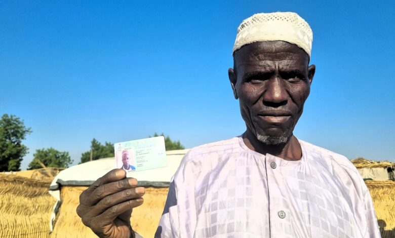 Man in traditional attire holding an ID card with thatched huts in the background under blue sky.