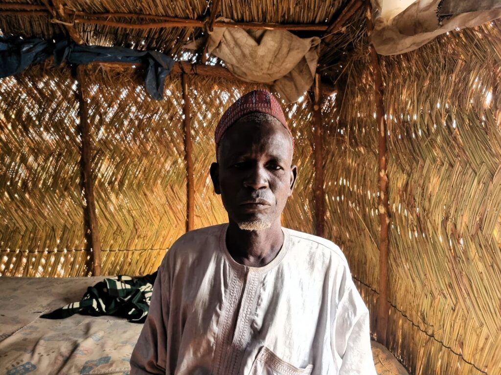 Elderly man with a cap posing inside a room with walls made of woven natural materials.