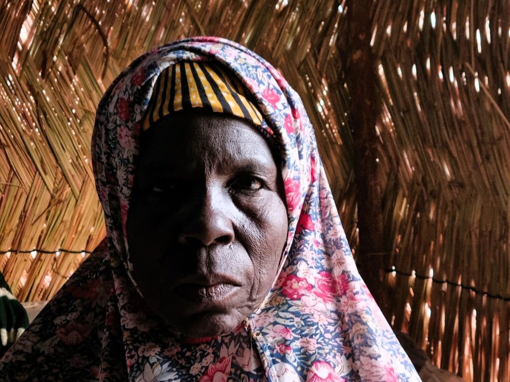 Elderly woman with a patterned headscarf in a hut with thatched walls.