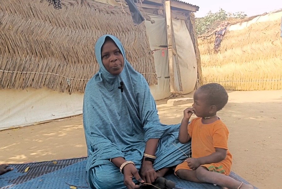 Woman in blue headscarf sitting with a young child in orange shirt, beside a thatched hut.