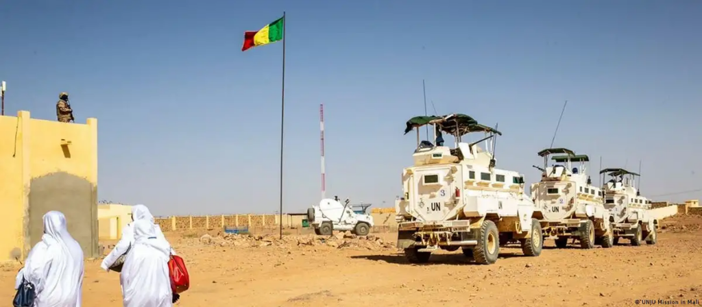 Desert checkpoint with UN vehicles, people in traditional dress, and a soldier under a flagpole.