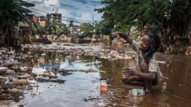 Girl squatting in polluted water, examining a bottle with murky liquid, surrounded by trash.
