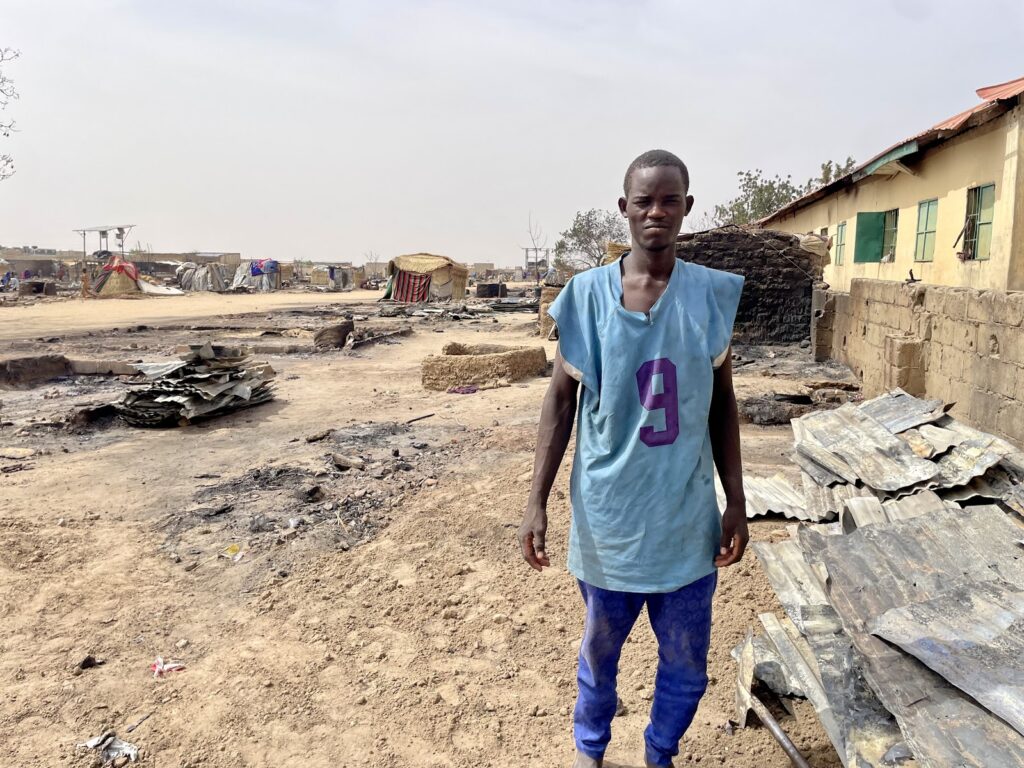 Young man stands in front of a burned and devastated settlement under a hazy sky.