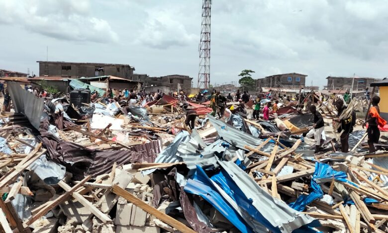 A devastated area with scattered debris, collapsed structures, and people amidst the ruins under a cloudy sky.