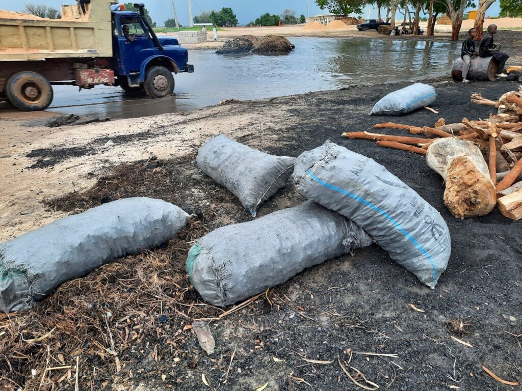 Sandbags on wet ground with a blue truck and two people sitting by the water in the background.