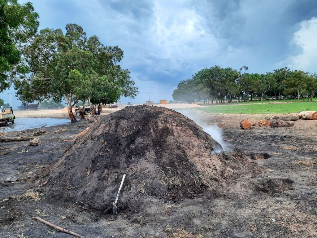 Traditional charcoal production mound emitting smoke with people working in the background under trees.